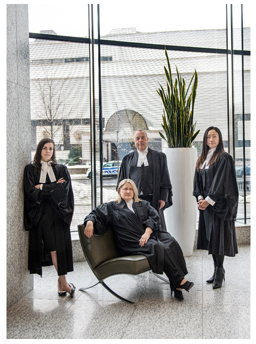 La photographie illustre quatre avocats vêtus de leurs robes officielles de la Cour de l’Ontario. Deux femmes et un homme sont debout, et une femme est assise sur un banc devant eux. Les quatre avocats posent, regardant vers l’appareil photo. Ils ont tous l’air sérieux. Ils se trouvent devant une grande fenêtre vitrée et d’un grand vase décoratif blanc contenant une plante tropicale verte. À l’arrière-plan, à l’extérieur, on aperçoit la façade du palais de justice d’Ottawa et une voiture de police d’Ottawa.